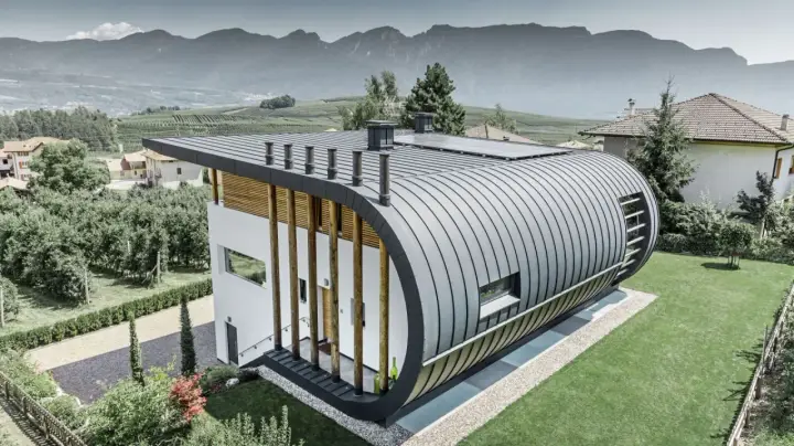 Nowoczesny dom z zaokrąglonym dachem z aluminium, które nie koroduje, otoczony zielenią i górami.